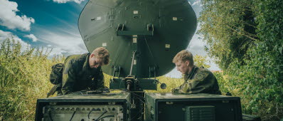Två soldater i uniform sitter på varsin sida om en rund satellitskiva som står ute i terrängen